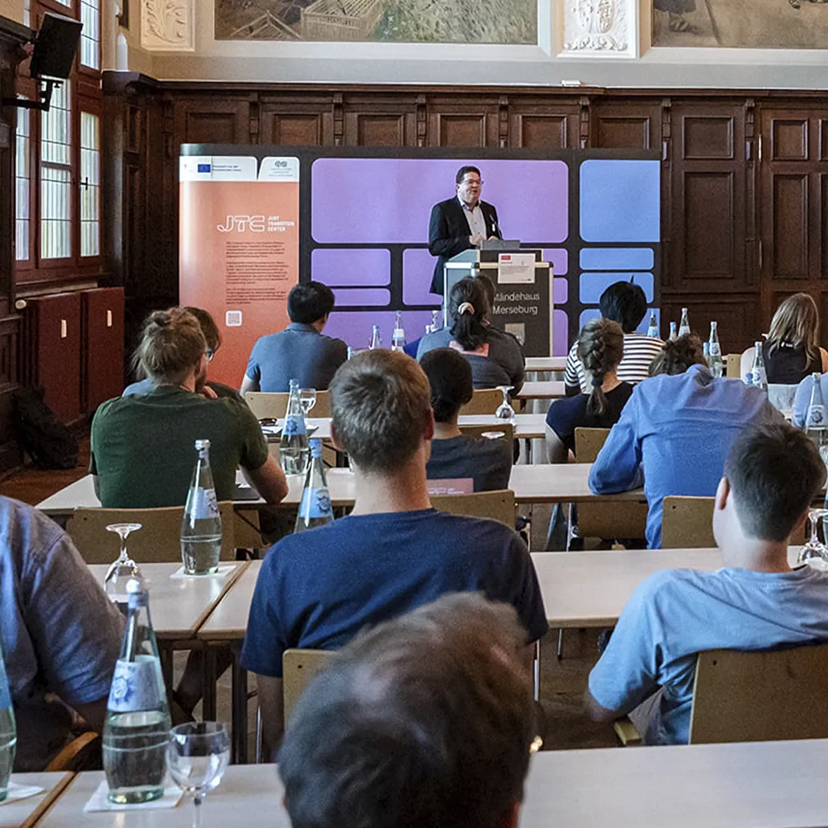 Foto einer Veranstaltung des JTC, eine Person hält eine Präsentation, im Hintergrund ist eine Messewand im Corporate Design des JTC zu sehen