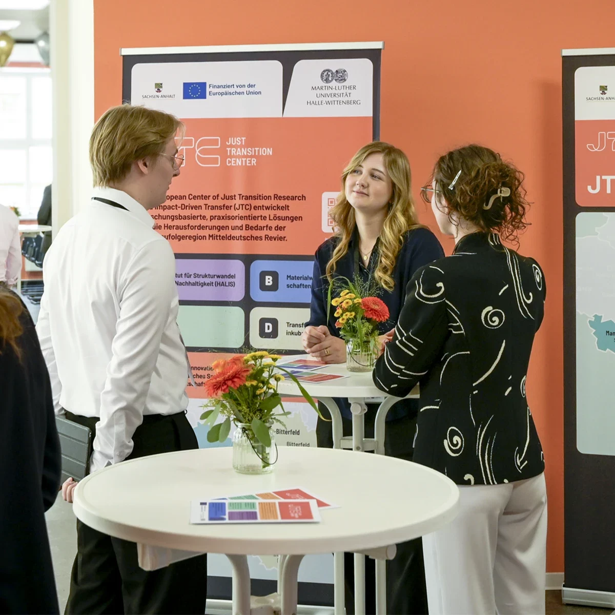 Foto einer Veranstaltung des JTC, drei Personen unterhalten sich an einem Stehtisch, im Hintergrund sind Rollups im Corporate Design des JTC zu sehen